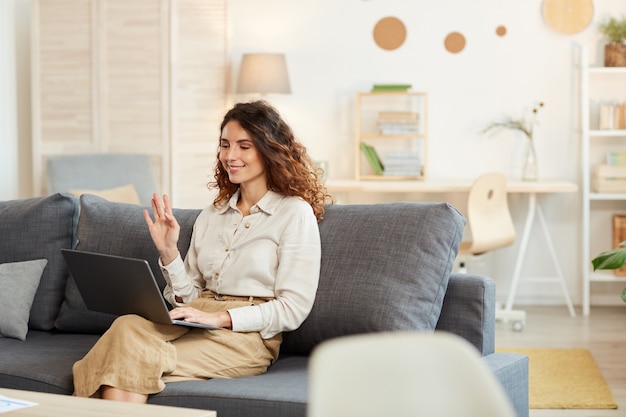 Jovem adulta sentada no sofá em casa, participando de uma videoconferência, cumprimentando seus colegas