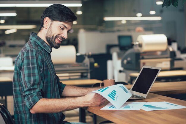 Jovem adulto alegre sentado à mesa do escritório com um laptop e uma caneca e sorrindo enquanto olha para os gráficos em suas mãos