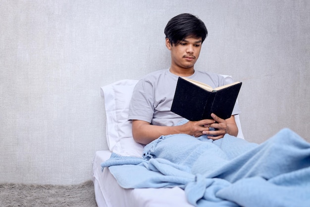 Jovem adulto asiático sentado na cama e lendo um livro antes de dormir