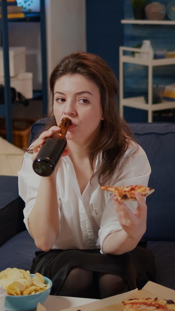 Jovem adulto bebendo cerveja de uma garrafa na sala de estar enquanto tem uma fatia de pizza na mão, sentado no sofá. mulher desfrutando de uma refeição com bebida alcoólica e fast food em um restaurante para viagem