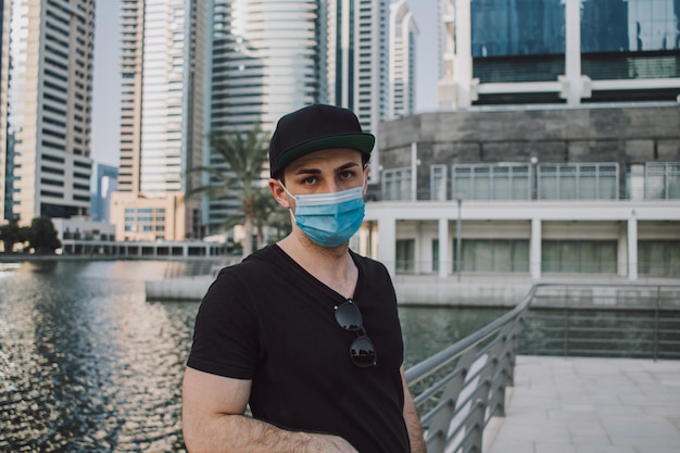 Foto jovem adulto com máscara médica em pé ao ar livre e olhando para a câmera com fundo de horizonte urbano da cidade