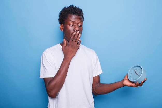 Jovem adulto de etnia afro-americana olha para a hora no relógio e cobre a boca em choque indivíduo masculino preto que está sempre a tempo carregando um relógio de parede e parecendo ansioso