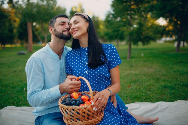 Jovem adulto mulher e homem casal sentado com cesta de frutas no prado de grama verde no parque. Piquenique de verão na natureza.