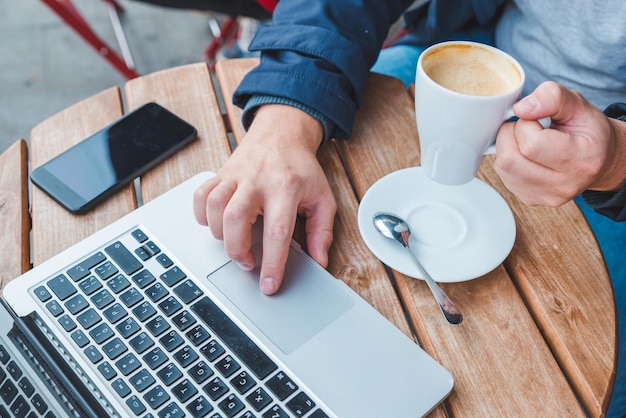 Jovem adulto trabalhando no laptop no café ao ar livre com xícara de café com leite