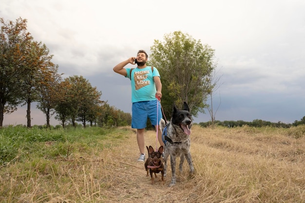 jovem adulto usando seu telefone inteligente enquanto passeava com seus cães ao pôr do sol no parque