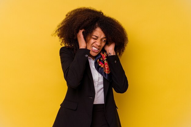 Jovem aeromoça afro-americana isolada em fundo amarelo, cobrindo as orelhas com as mãos.