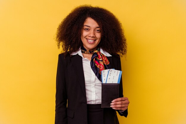 Jovem aeromoça afro-americana segurando uma passagem de avião isolada em amarelo feliz, sorridente e alegre.