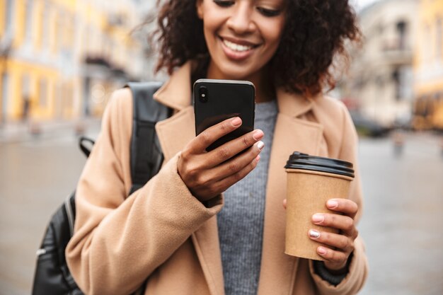 Jovem africana alegre, vestindo um casaco, caminhando ao ar livre, segurando uma xícara de café para viagem, usando um telefone celular