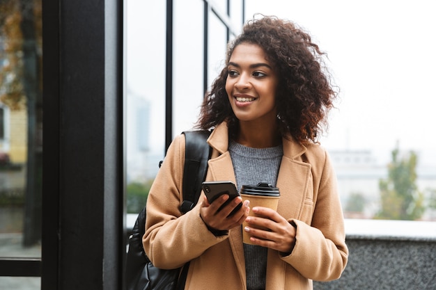 Jovem africana alegre, vestindo um casaco, caminhando ao ar livre, segurando uma xícara de café para viagem, usando um telefone celular