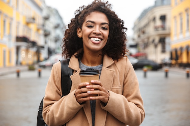 Jovem africana alegre, vestindo um casaco e caminhando ao ar livre, segurando uma xícara de café para viagem