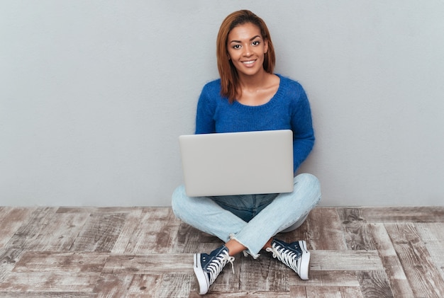 Jovem africana de suéter e jeans sentada no chão de madeira com o laptop