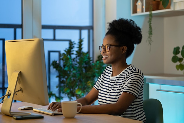 Jovem africana feliz trabalhadora remota usando óculos digitando no computador desfrutando de trabalho freelance