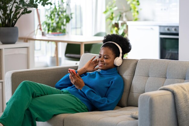 Foto jovem africana relaxada com fones de ouvido segurando telefone ouvindo música favorita ou podcast em casa