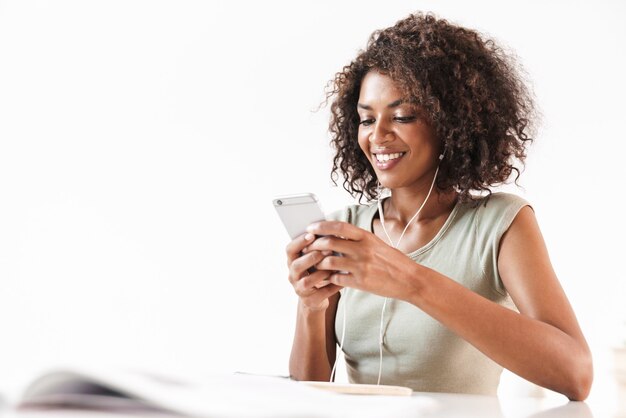 Jovem africana sorridente, sentada à mesa isolada na parede branca, ouvindo música com fones de ouvido, segurando o telefone celular