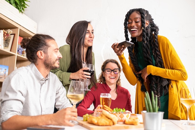 Jovem africana sorrindo enquanto está sentada com seus amigos em um bistrô tirando fotos de sua refeição com seu influenciador de smartphone desfrutando de comida e bebida