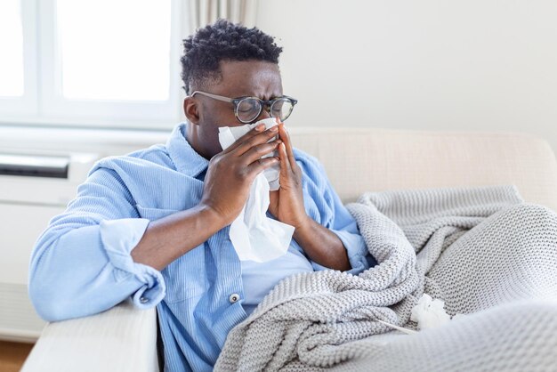 jovem africano coberto com um cobertor soprando nariz correndo pegou febre apanhou frio espirrando em tecido sentar-se no sofá homem preto alérgico doente com sintomas de alergia tossindo em casa corona 19