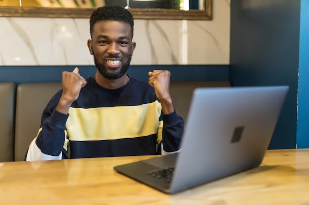 Jovem africano com laptop comemorando em uma cafeteria moderna