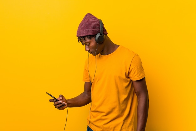 Jovem Africano de pé contra um amarelo usando um chapéu, ouvindo música com um telefone