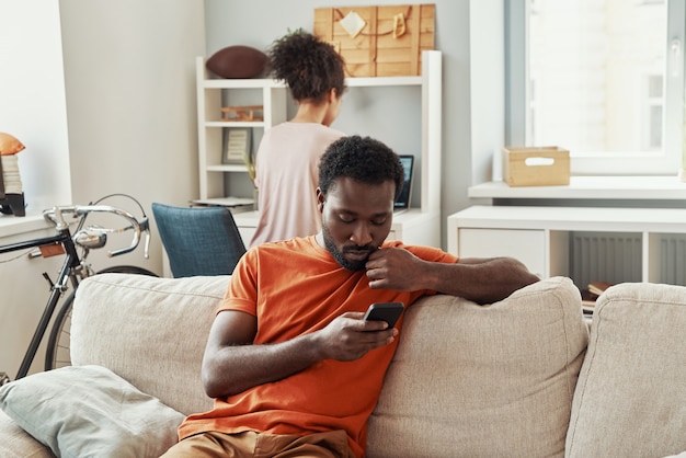 Jovem africano despreocupado usando um smartphone enquanto passa um tempo em casa com a namorada