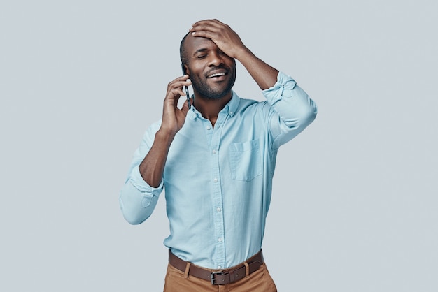 Foto jovem africano feliz falando no telefone inteligente e sorrindo enquanto fica de pé contra uma parede cinza