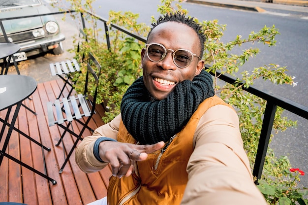 Jovem africano feliz sorrindo ao ar livre tira uma selfie fazendo sinal de v com a mão