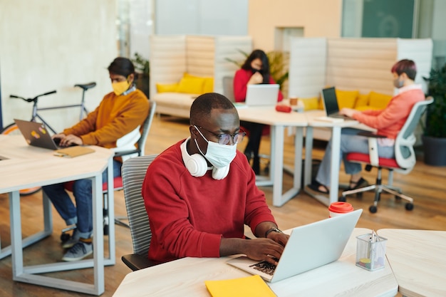 Jovem africano gerente de escritório em trajes casuais e máscara protetora usando laptop enquanto prepara a apresentação contra colegas