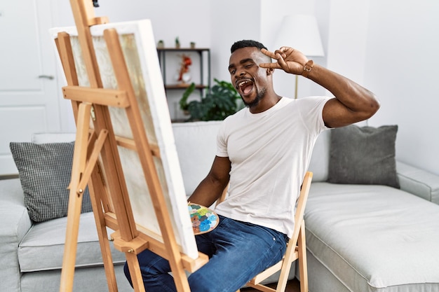 Jovem africano pintando sobre tela em casa fazendo o símbolo da paz com os dedos sobre o rosto, sorrindo alegre mostrando vitória