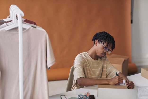 Jovem africano sentado à mesa preenchendo o formulário no pacote antes de enviar ele trabalhando no depósito