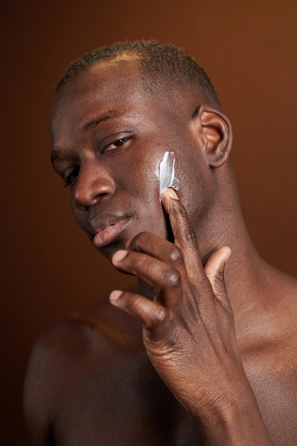 Jovem africano sério aplicando creme hidratante no rosto