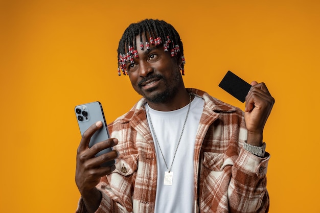 Jovem africano sorridente em fundo amarelo usando telefone celular e segurando cartão de crédito