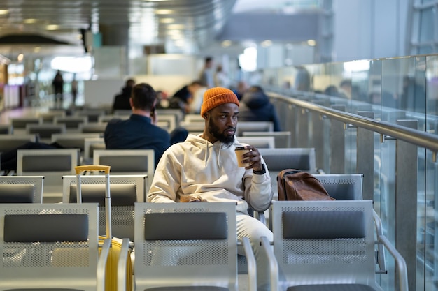 Jovem africano tomando café e comendo sanduíche enquanto espera para voar no terminal do aeroporto