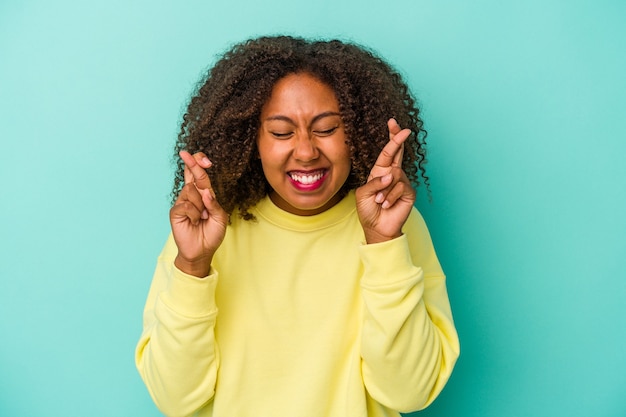 Jovem afro-americana com cabelo encaracolado isolado em um fundo azul cruzando os dedos para ter sorte