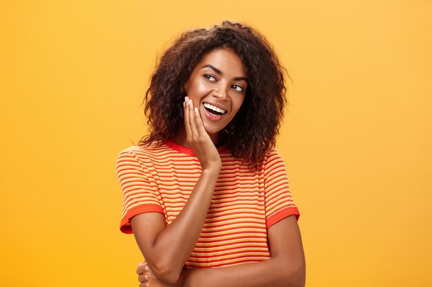 Jovem afro-americana com camiseta listrada, inclinando a cabeça na palma da mão e olhando com expressão curiosa