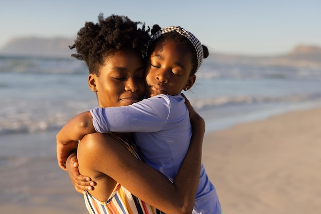 Jovem afro-americana com os olhos fechados, abraçando a filha contra o mar e o céu claro ao pôr do sol