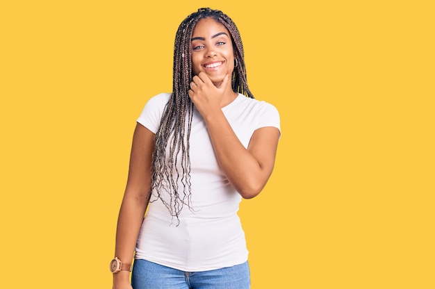 Jovem afro-americana com tranças vestindo roupas casuais parecendo confiante para a câmera sorrindo com braços cruzados e mão levantada no queixo pensando positivo