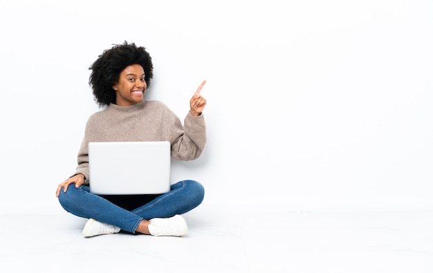 Jovem afro-americana com um laptop sentada no chão apontando o dedo para o lado