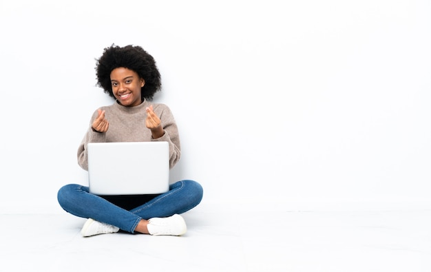 Jovem afro-americana com um laptop sentada no chão fazendo gestos de dinheiro