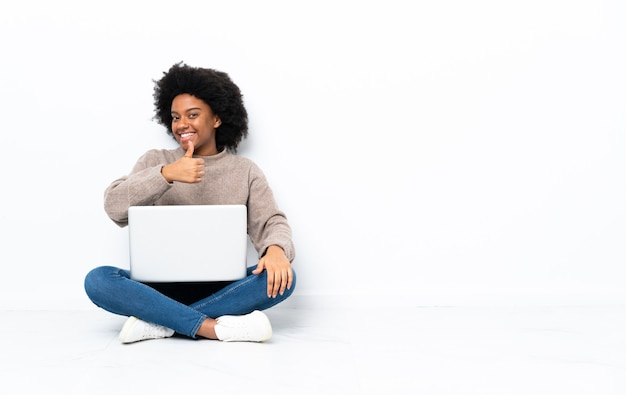 Jovem afro-americana com um laptop sentada no chão fazendo um gesto de polegar para cima