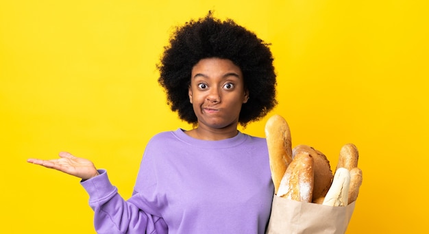Jovem afro-americana comprando algo, pão isolado em amarelo, tendo dúvidas ao levantar as mãos