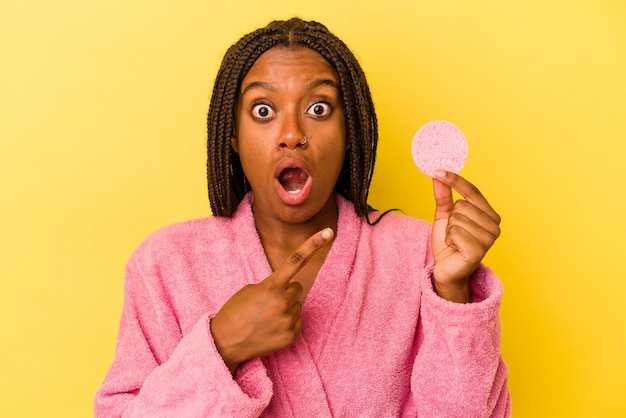 Jovem afro-americana de roupão segurando um disco de maquiagem isolado em um fundo amarelo apontando para o lado