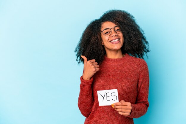 Jovem afro-americana encaracolada segurando um cartaz com um sim, sorrindo e levantando o polegar