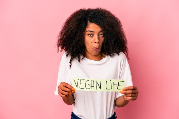 Jovem afro-americana encaracolada segurando um cartaz de vida vegana encolhe os ombros e abre os olhos confusos.