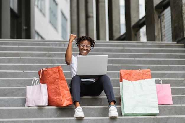 Jovem afro-americana fazendo compras online usando laptop sentado nos degraus do shopping