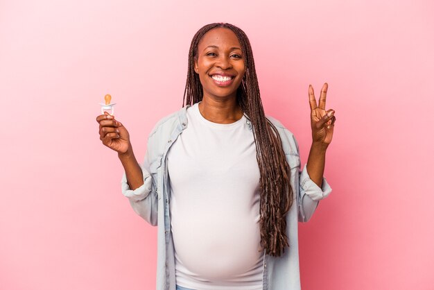 Jovem afro-americana grávida segurando a chupeta isolada no fundo rosa, mostrando o número dois com os dedos.