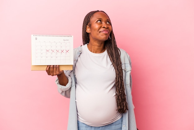 Jovem afro-americana grávida segurando calendário isolado no fundo rosa, sonhando em alcançar objetivos e propósitos