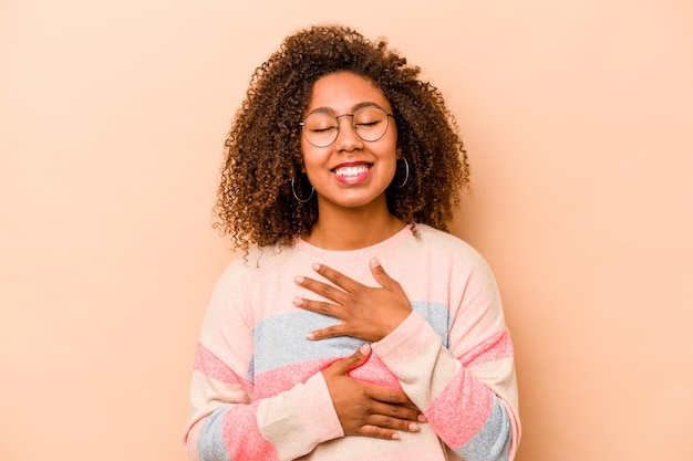 Jovem afro-americana isolada em fundo bege ri alegremente e se diverte mantendo as mãos no estômago