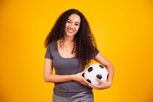 Jovem afro-americana isolada em um fundo amarelo com uma bola de futebol