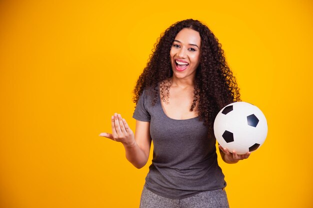 Jovem afro-americana isolada em um fundo amarelo com uma bola de futebol