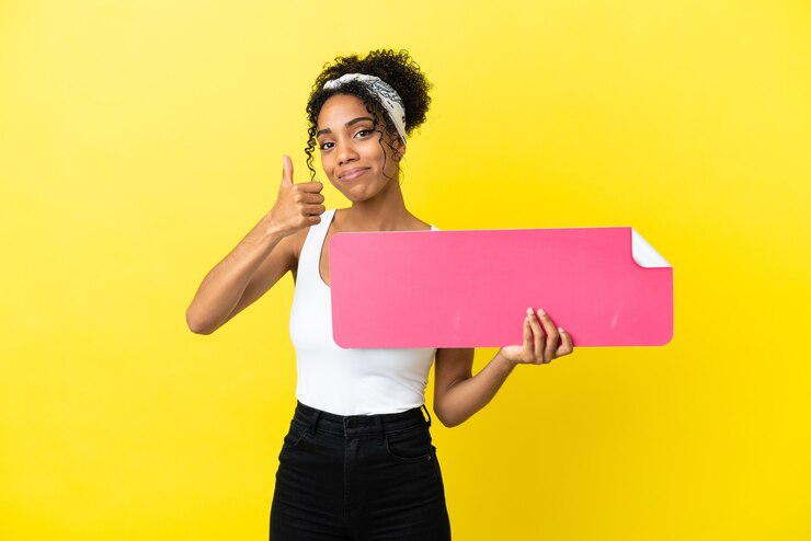 Foto jovem afro-americana isolada em um fundo amarelo segurando um cartaz vazio com o polegar para cima