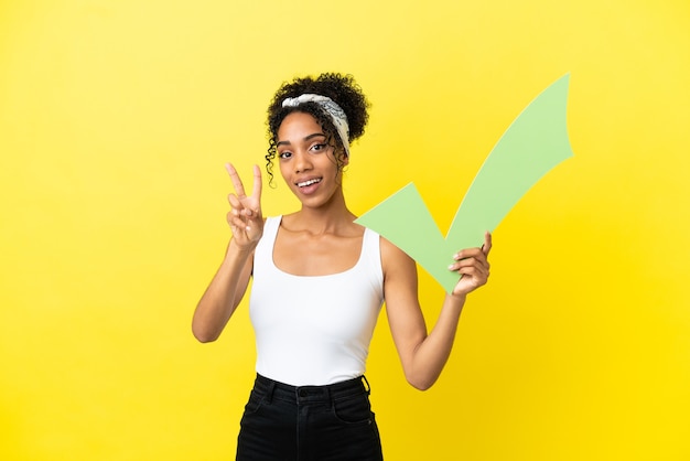 Jovem afro-americana isolada em um fundo amarelo segurando um ícone de seleção e comemorando uma vitória